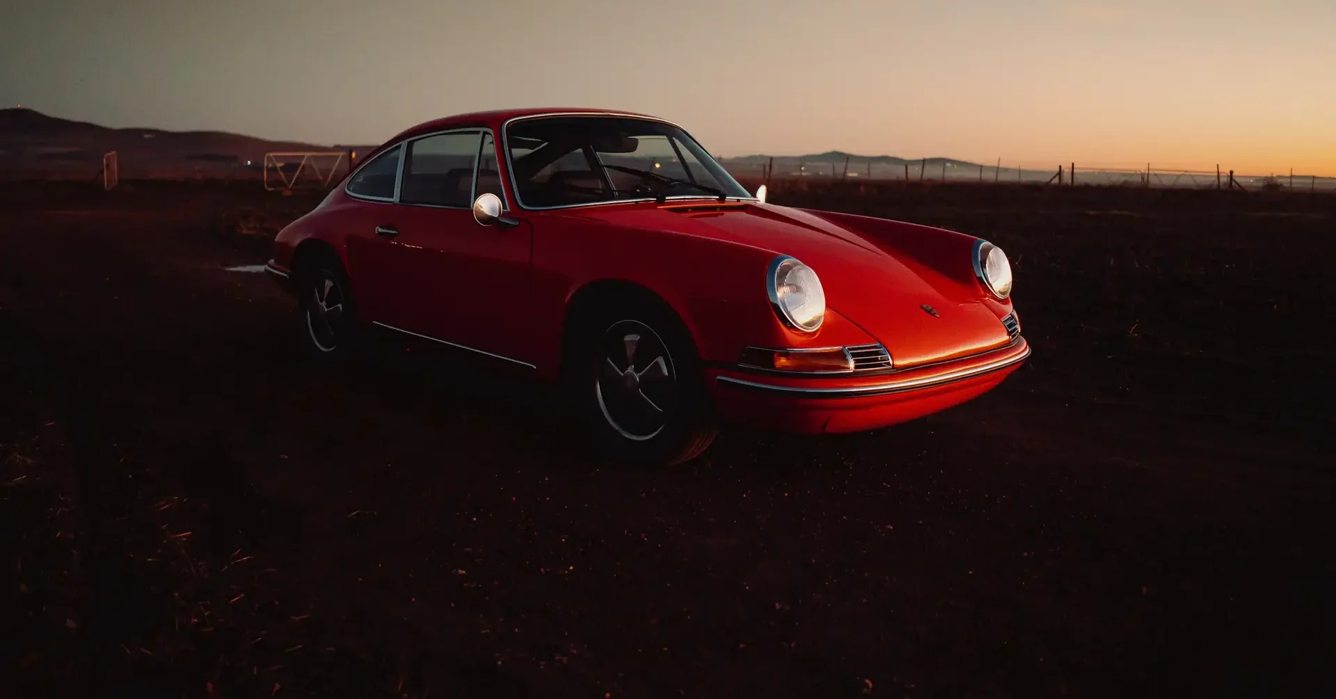 Aces Auto Elite - A classic red Porsche 911 coupe parked on a dirt road at sunset, with mountains and a fence in the background under a clear sky. | Arizona Luxury & Exotic Car Service