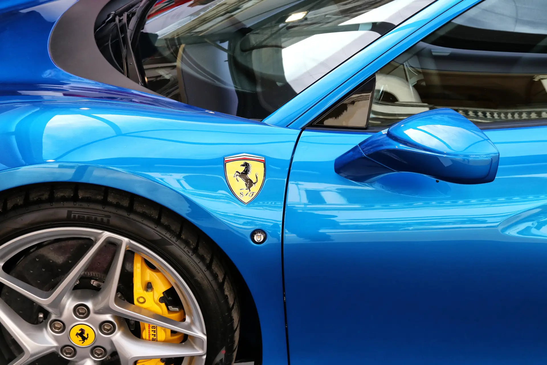 Aces Auto Elite - Close-up of a shiny blue Ferrari sports car showing the door, side mirror, and part of the front wheel with yellow brake calipers. The Ferrari logo is visible on the side above the wheel. | Arizona Luxury & Exotic Car Service