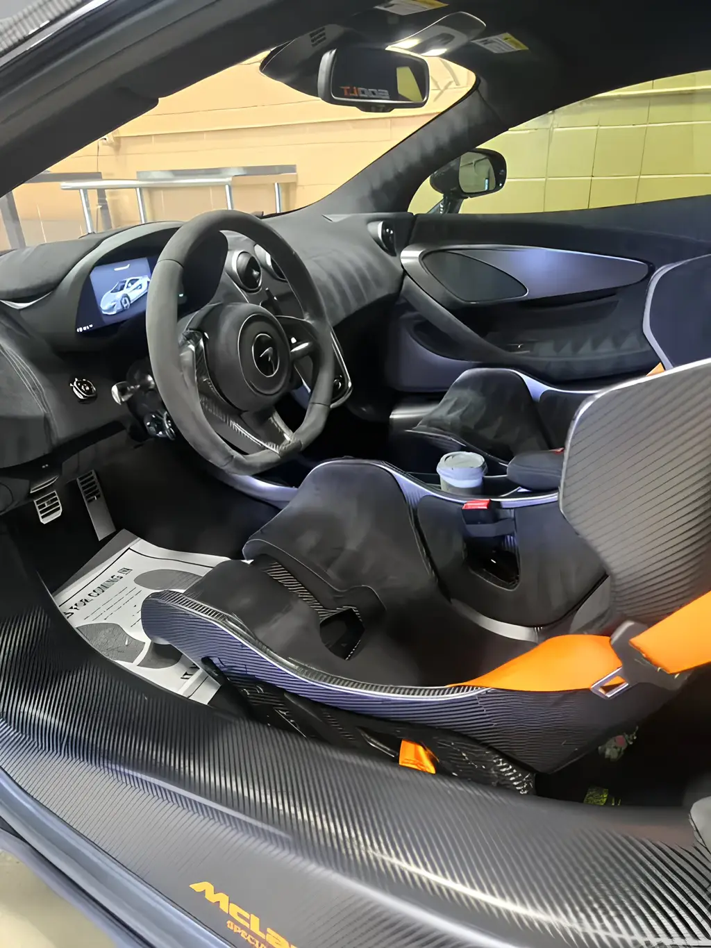 Aces Auto Elite - Interior view of a McLaren sports car, showing the drivers seat with orange seatbelt, carbon fiber accents, steering wheel, digital dashboard display, and a cup in the center console. | Arizona Luxury & Exotic Car Service