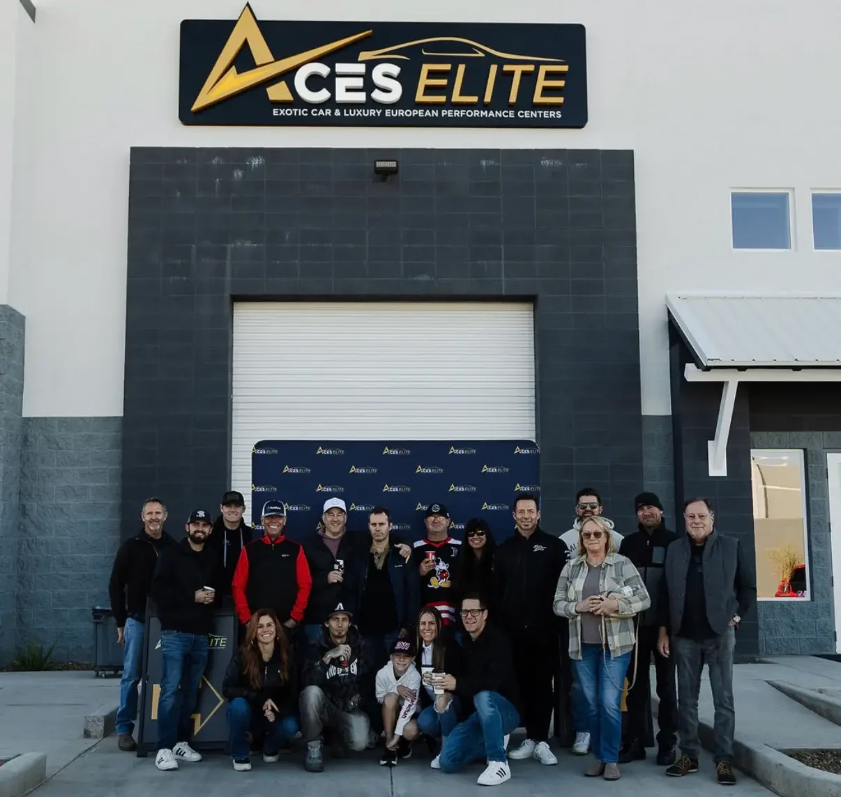 Aces Auto Elite - A group of people pose together for a photo outside a building with a sign that reads Aces Elite Exotic Car & Luxury European Performance Centers. The group stands on a concrete driveway in front of a large closed garage door. | Arizona Luxury & Exotic Car Service