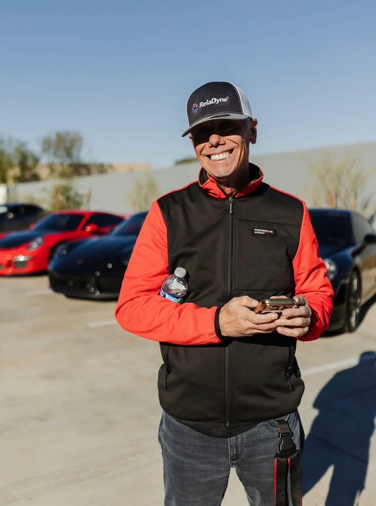 Aces Auto Elite - A man wearing a black and red jacket and a RelaDyne cap stands smiling in a parking lot, holding a phone and water bottle, with several sports cars parked behind him on a sunny day. | Arizona Luxury & Exotic Car Service
