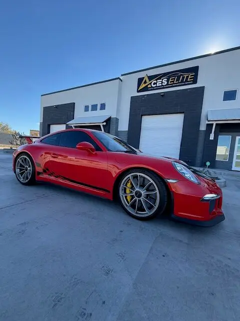 Aces Auto Elite - A red Porsche sports car with black detailing is parked in front of a modern building with a sign that reads Aces Elite under a clear blue sky. | Arizona Luxury & Exotic Car Service