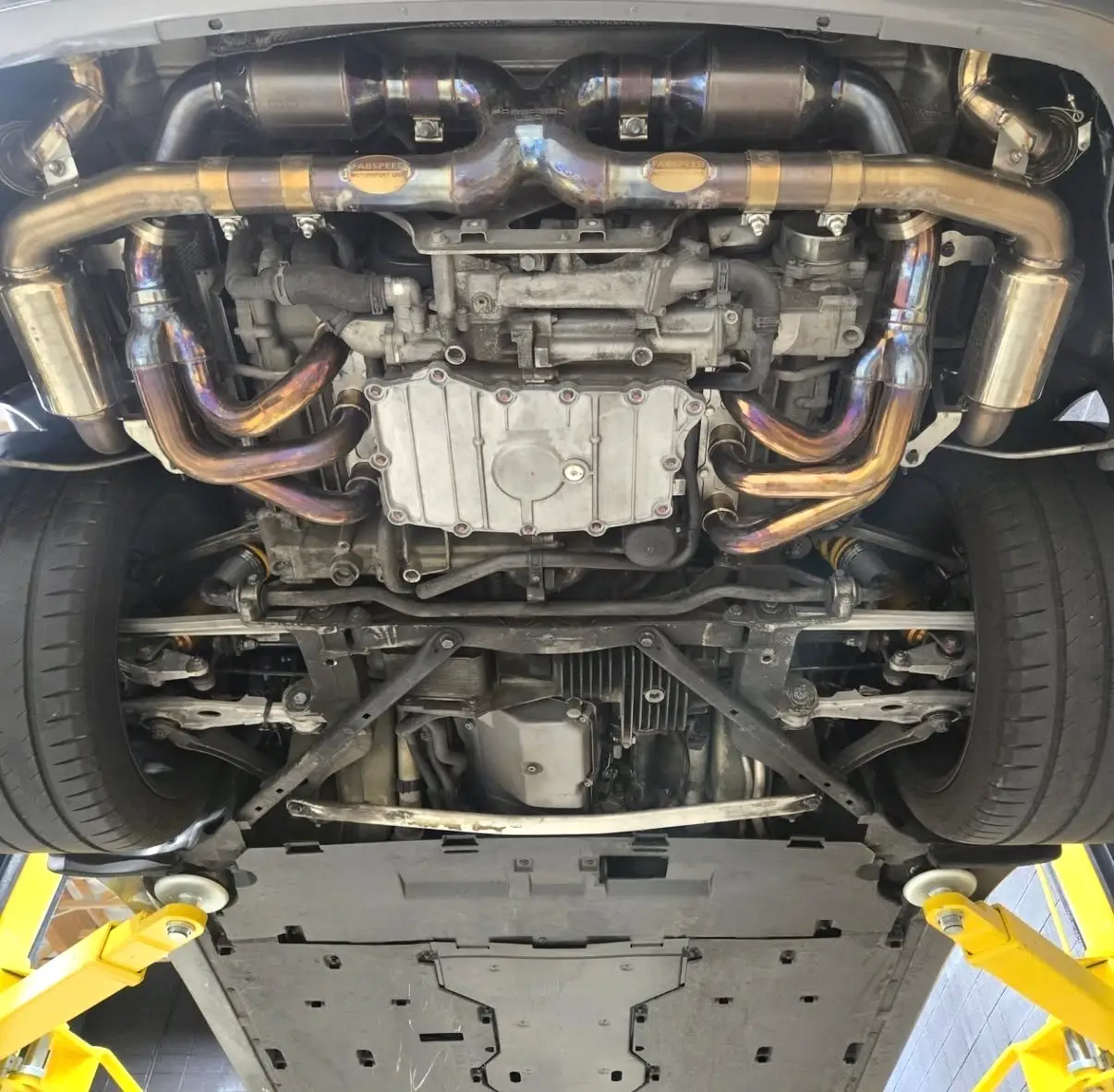 Aces Auto Elite - View of a car’s undercarriage from below, showing exposed exhaust system, engine, and suspension components, with the car lifted on yellow hydraulic ramps. | Arizona Luxury & Exotic Car Service
