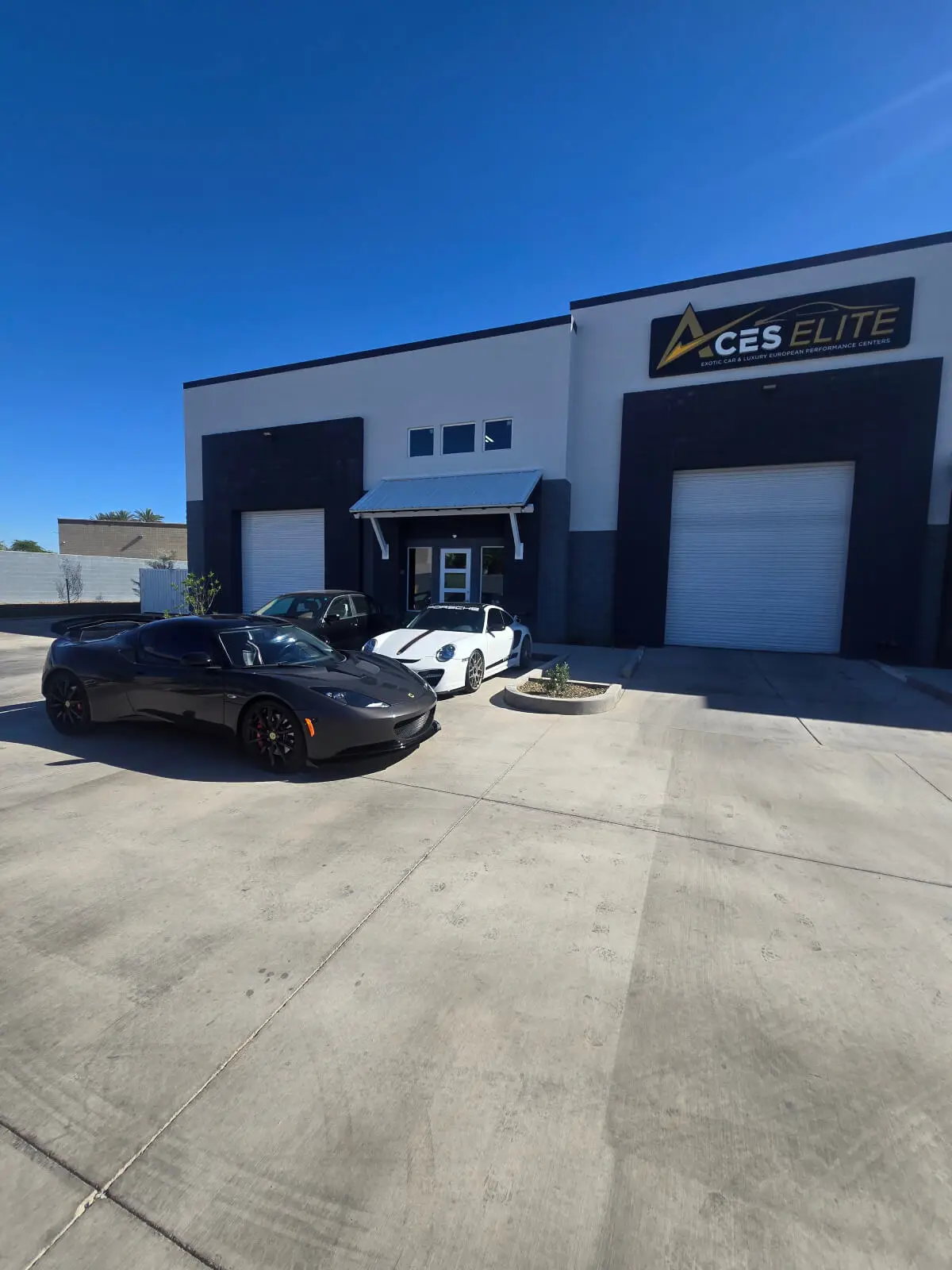 Aces Auto Elite - Two sports cars, one black and one white, are parked outside a modern commercial building with a sign that reads Aces Elite, hinting at the founder’s passion for excellence. The sky above is clear and blue. | Arizona Luxury & Exotic Car Service