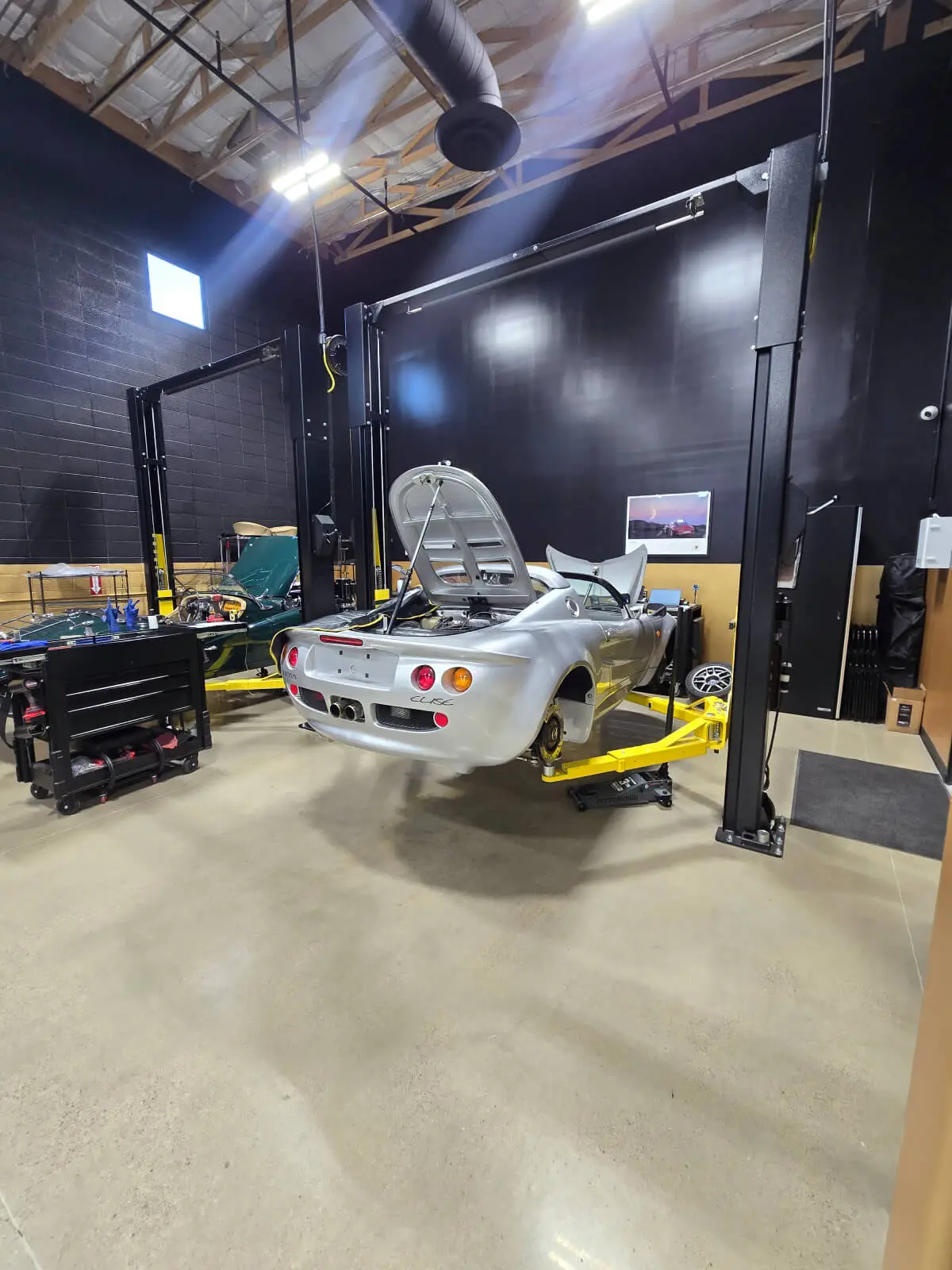 Aces Auto Elite - A silver sports car with its hood open is elevated on a hydraulic lift inside a spacious, well-lit auto repair shop with tools and equipment visible nearby. | Arizona Luxury & Exotic Car Service
