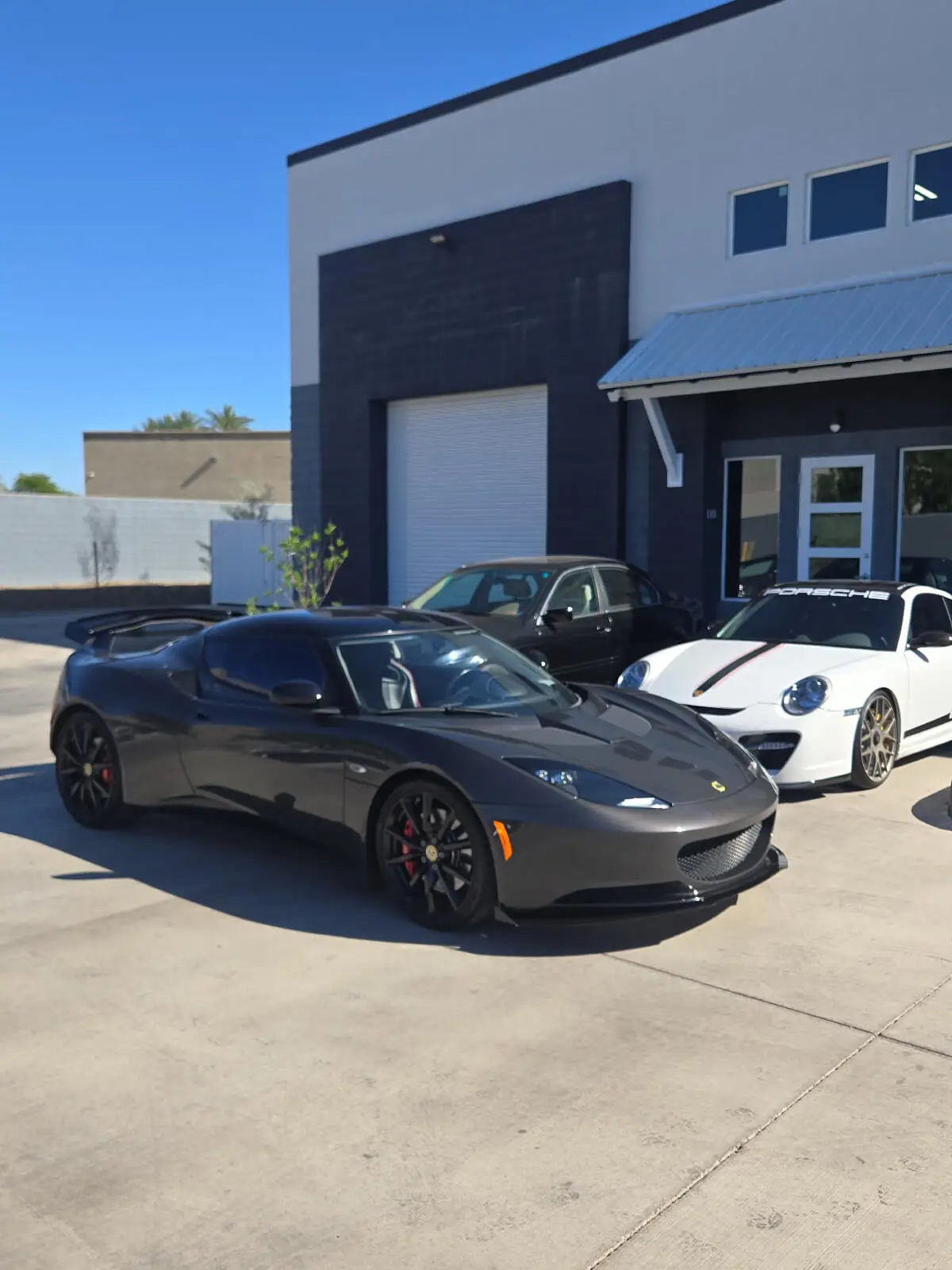 Aces Auto Elite - A dark gray sports car is parked in front of a white commercial building, alongside a white Porsche and a black car, all under a clear blue sky. | Arizona Luxury & Exotic Car Service