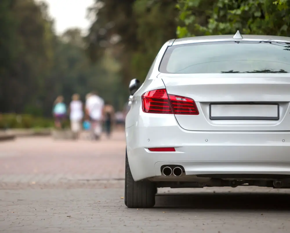 Aces Auto Elite - A silver car is parked on a paved street next to greenery, with four blurry people walking in the background. The cars rear and taillights are visible, and the license plate is blank. | Arizona Luxury & Exotic Car Service