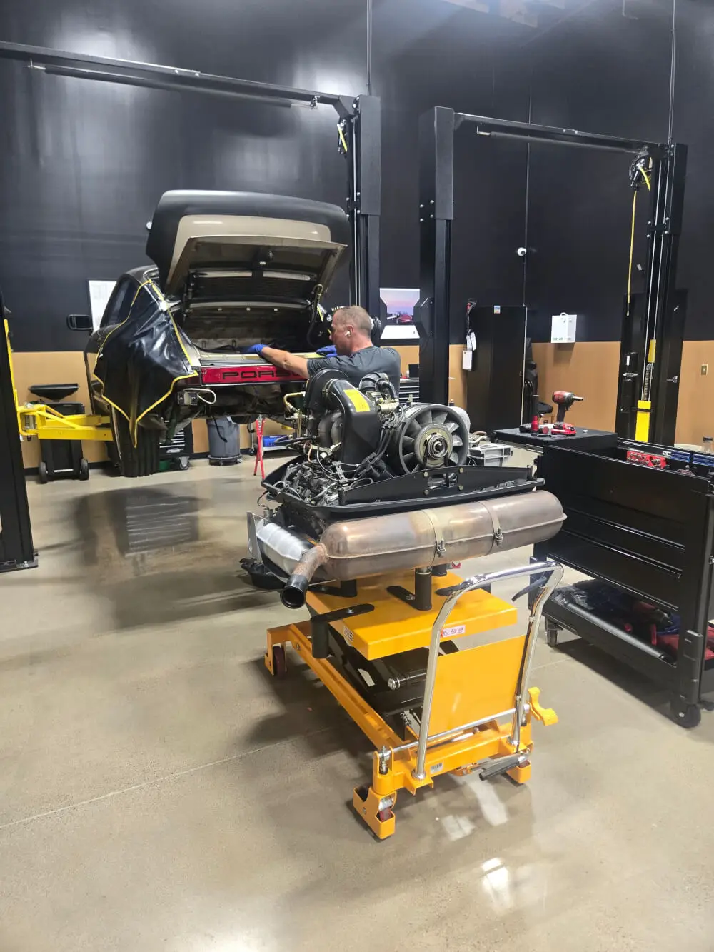 Aces Auto Elite - A mechanic works on a car with its trunk open in a brightly lit garage. An engine is mounted on a yellow platform nearby, ready to be installed or removed. Tools and equipment surround the workspace. | Arizona Luxury & Exotic Car Service