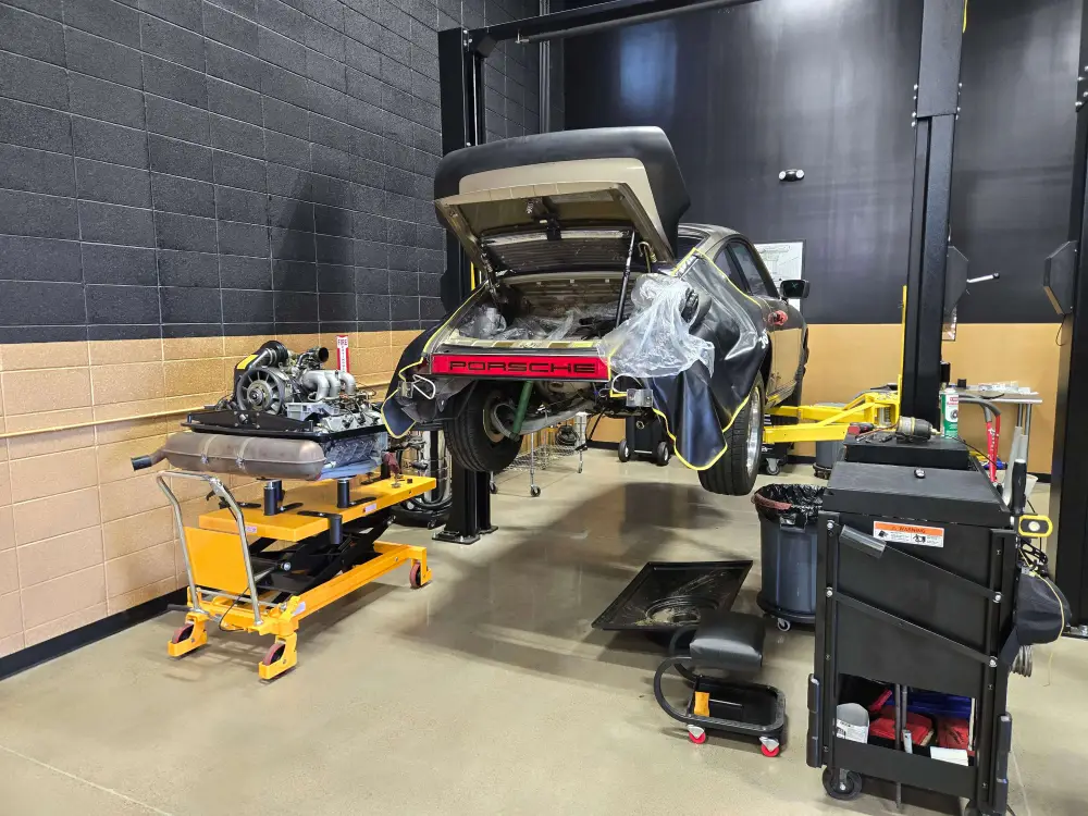 Aces Auto Elite - A Porsche car is elevated on a hydraulic lift in a garage, rear open and engine removed for performance upgrades. The engine rests on a yellow stand, while tools and equipment are organized around the workspace for seamless customization. | Arizona Luxury & Exotic Car Service