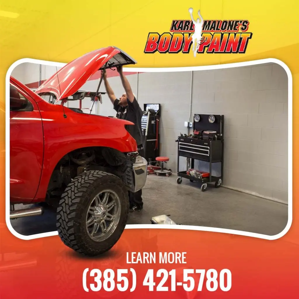 An auto technician lifting the hood of a red pickup truck undergoing front-end structural body repair inside a shop bay.