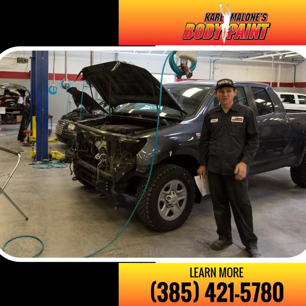 An auto body technician holding paperwork in front of a dark grey pickup truck with its front grille and bumper disassembled in a service bay.