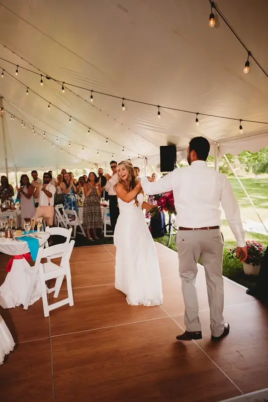 Wedding couple dancing on a wood dance floor under a large event tent with string lights and guests