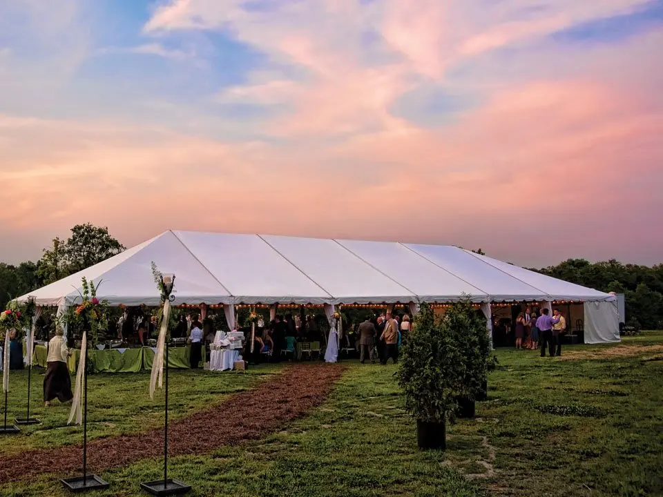 Large white wedding tent set up on a grassy outdoor venue at sunset with guests gathered inside