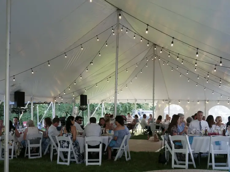 A lively outdoor event under a white tent adorned with string lights. Guests sit at round tables with white chairs, enjoying a festive, cheerful atmosphere.