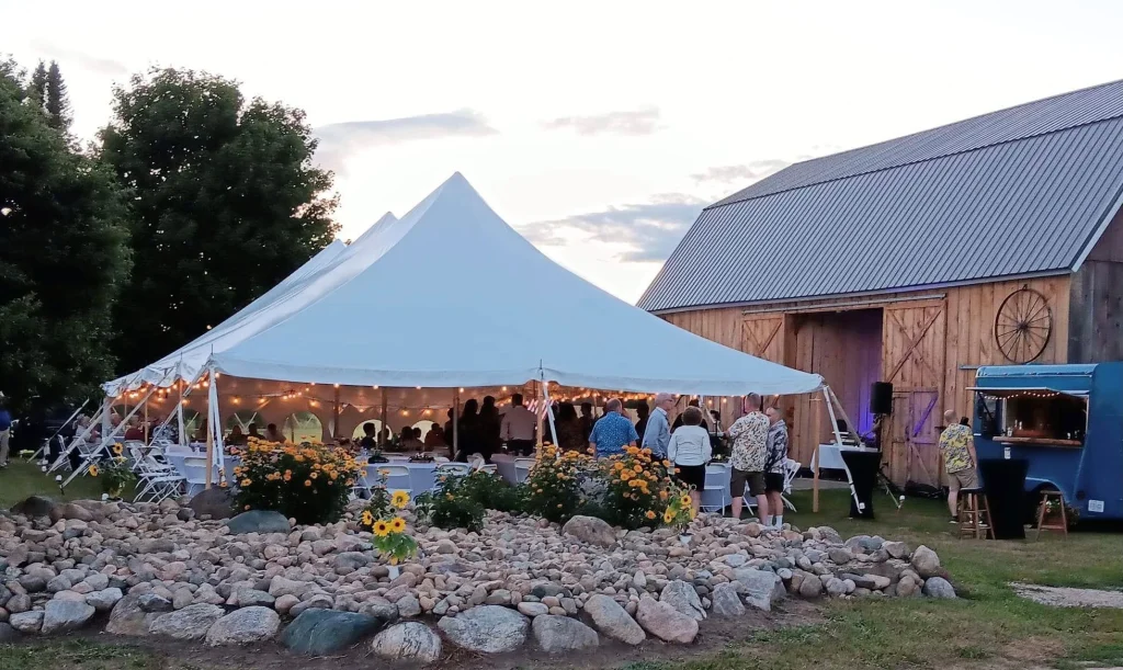 A white tent with string lights hosts a gathering beside a rustic barn at sunset. People mingle around as flowers and rocks border the cheerful scene.