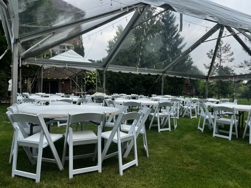 Backyard event tent rentals featuring a clear frame tent with tables, white chairs, and string lighting prepared for an outdoor celebration.