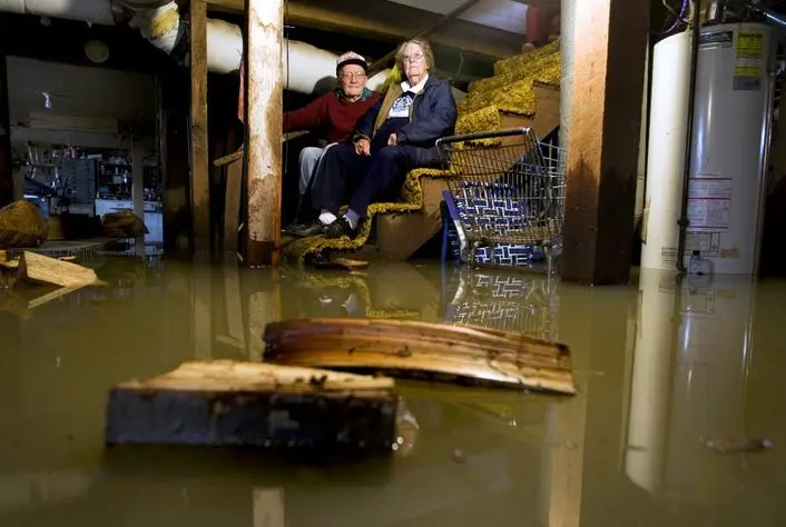Elderly couple sitting on stairs in flooded basement, surrounded by water and debris, highlighting the need for basement waterproofing solutions in Hopewell.