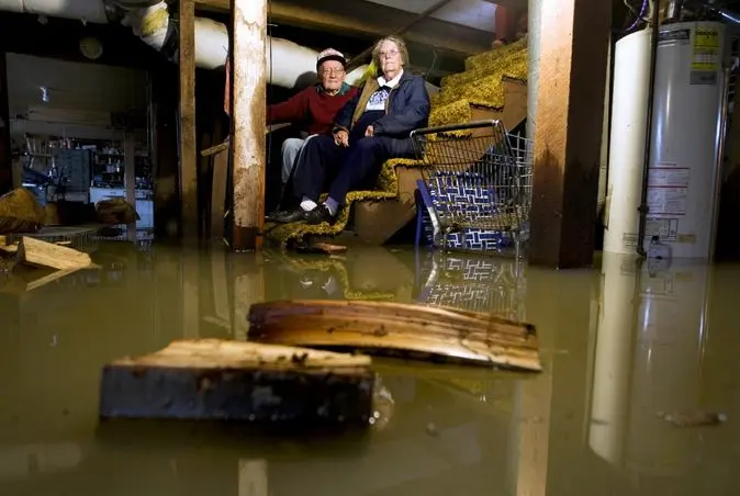 Elderly couple sitting on stairs in flooded basement, surrounded by water and debris, highlighting the need for basement waterproofing solutions in Hopewell.