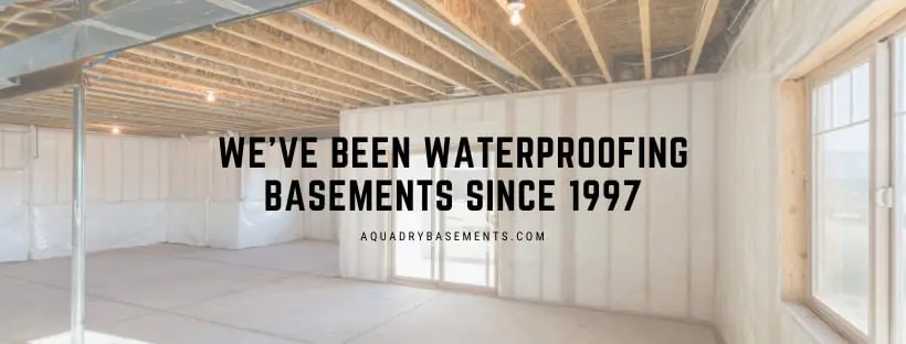 Interior view of a waterproofed basement with white walls and exposed wooden beams, featuring the text "We've been waterproofing basements since 1997" and the website "aquadrybasements.com," emphasizing AquaDry's expertise in basement waterproofing services.