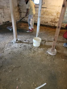Man sweeping a dirt floor in a basement with a shovel, near wooden beams and a white bucket. Exposed brick walls create a rugged, unfinished look.