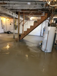 A basement with freshly poured concrete, wooden stairs, a water heater, and exposed utilities. The space is brightly lit, clean, and utilitarian.