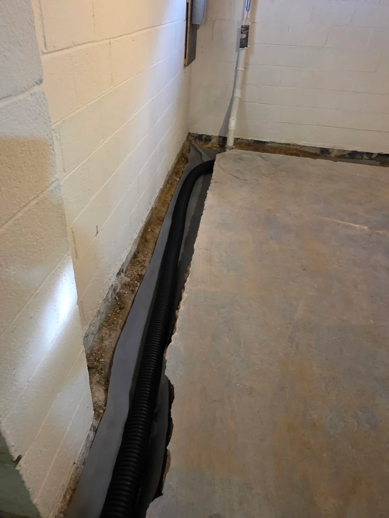 A basement corner features exposed concrete foundation with a drainage pipe installed along the wall base, suggesting recent waterproofing work.