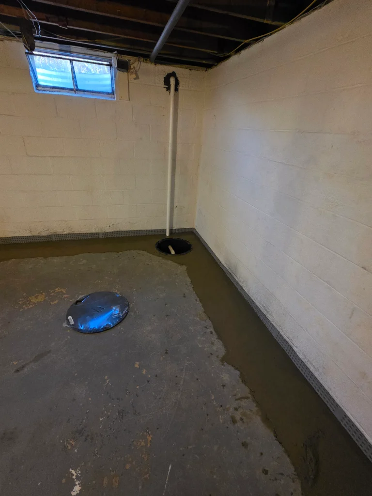 Basement with exposed beams and a small window. A sump pump and drainage system are visible along the floor perimeter. The room appears under renovation.