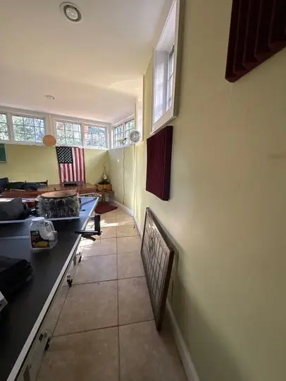A bright home office with green walls and an American flag hanging. Sunlight streams through large windows. Items are scattered, creating a cozy yet cluttered feel.