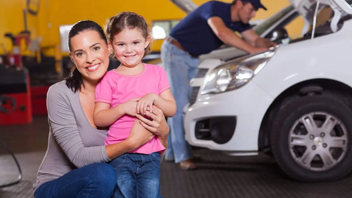 Smiling woman and young girl in an auto repair shop, with a mechanic working on a vehicle in the background, emphasizing family-friendly service and convenience at Deutmeyer Auto Advantage.
