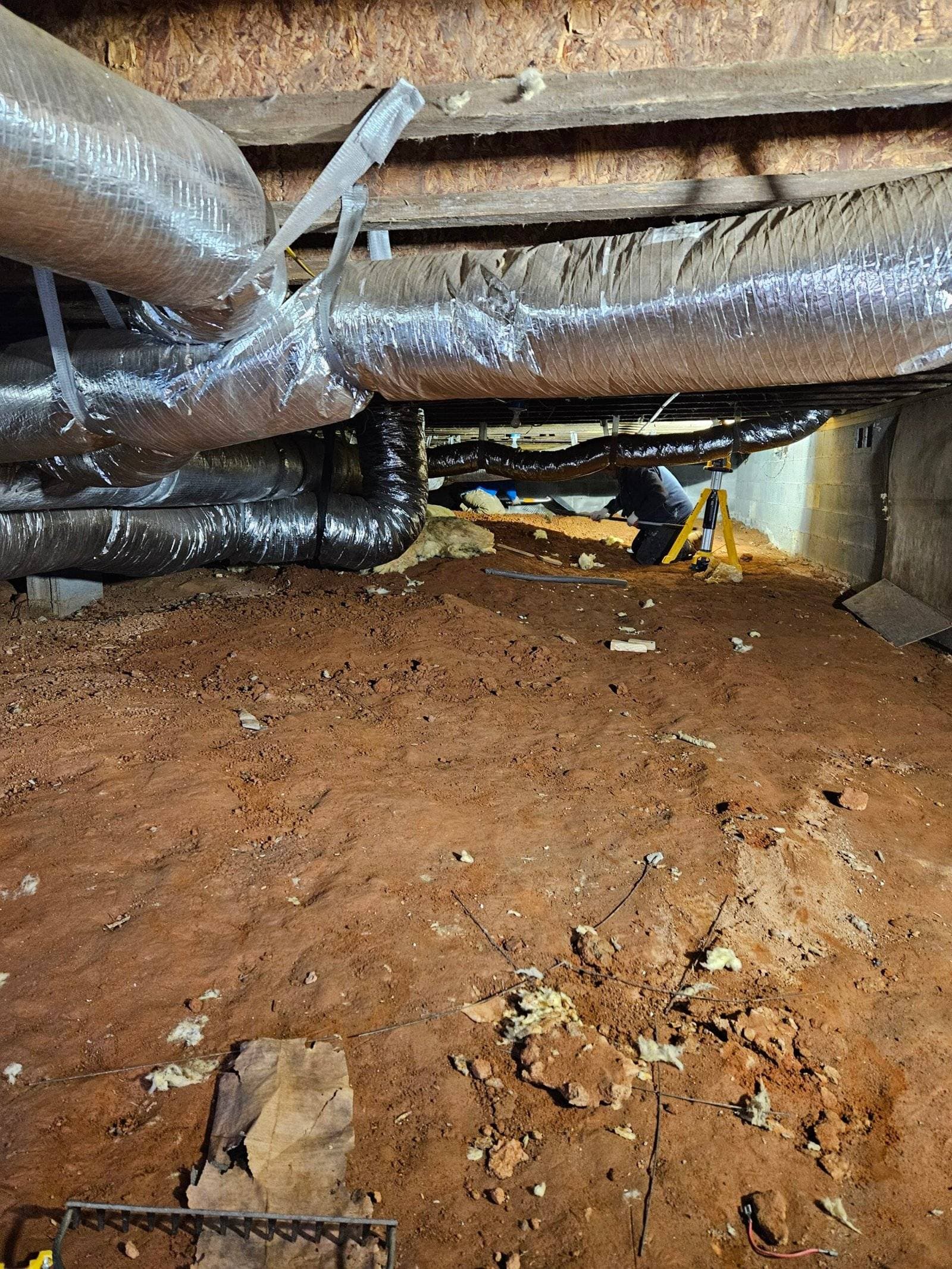 Crawl space with exposed dirt floor, visible ductwork, and restoration worker assessing moisture damage for water remediation services.