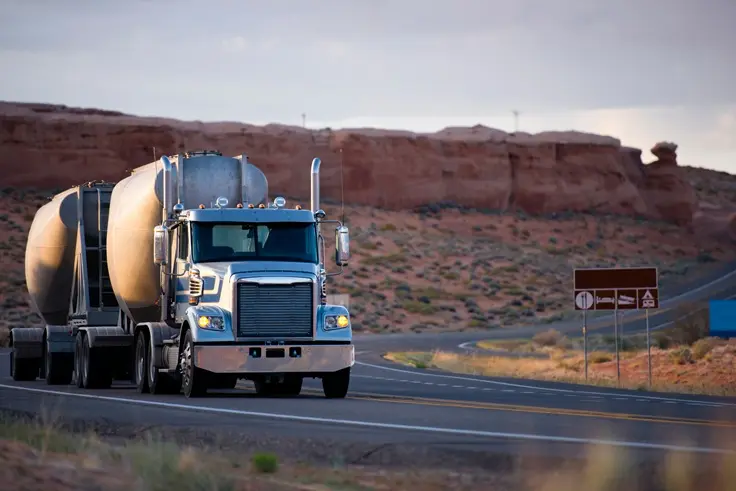 Big rig American classic semi truck with two compact bulk semi trailer going on winding road with red rocks hills of Arizona landscape in twilight with sunlight and shadow