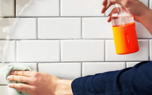 Person cleaning white wall tiles with spray bottle and towel