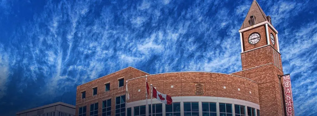 Canadian flags fly in front of a brick building with a clock tower under a dramatic blue sky. Takeoff-Moving Toronto
