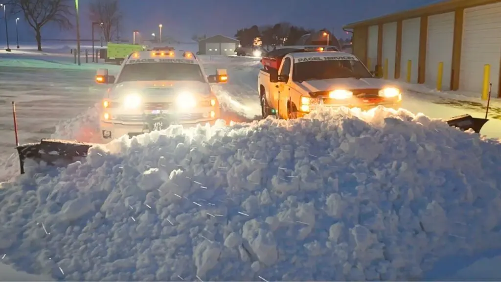 Two Pagel Services trucks with headlights on are shown plowing a large amount of snow in a commercial parking lot during early morning hours. Snow is being pushed into a central pile as snowflakes fall lightly.