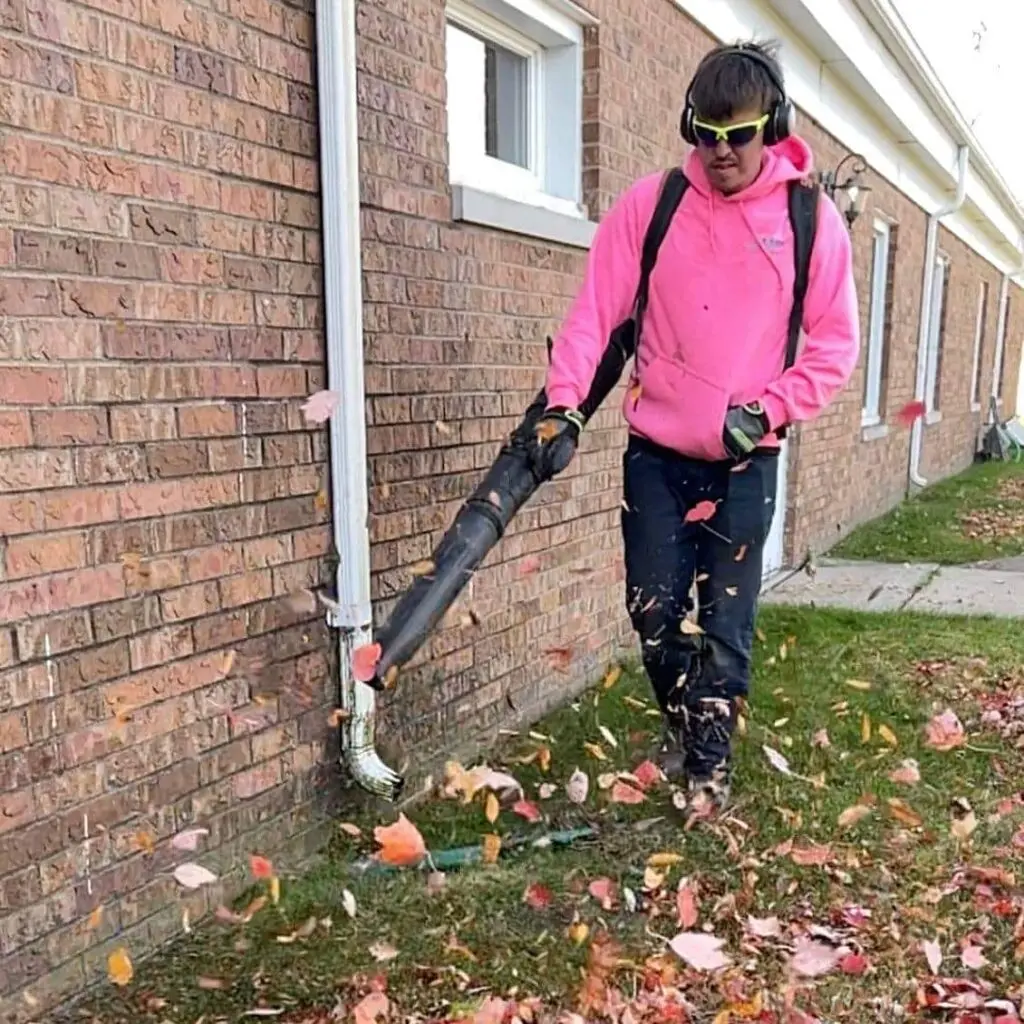 A worker wearing a bright pink hoodie, sunglasses, and ear protection uses a backpack leaf blower to clear fallen leaves from the edge of a brick building onto a grassy area during a fall clean-up.