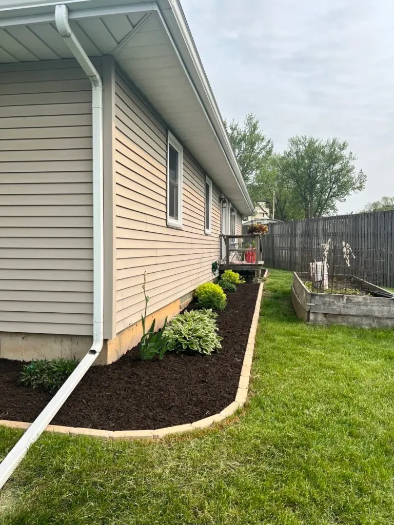A neatly edged landscape bed filled with dark brown mulch runs along the side of a beige house. Various green plants and hostas line the bed, which is bordered with light tan concrete edging.