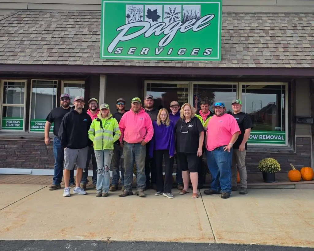 A group of 15 Pagel Services employees stands smiling outside their office building under a large green company sign. Some are wearing bright safety gear and pink sweatshirts, with pumpkins placed near the entrance.