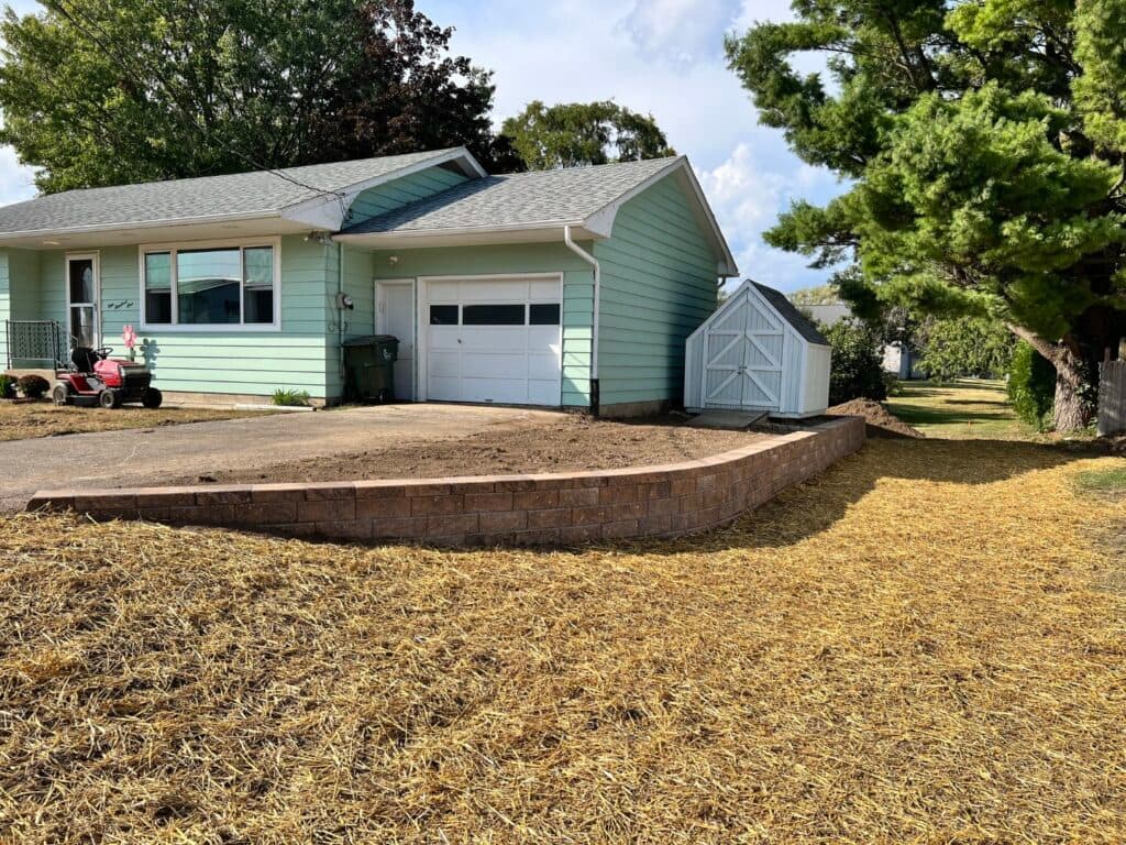 A freshly installed brown retaining wall wraps around the side of a light green house with a white garage. The yard is covered in straw, indicating recent grading and seeding as part of a landscaping project.