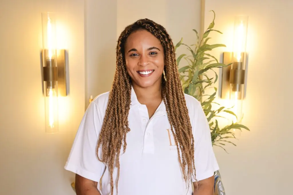 Smiling woman with long, braided hair wearing a white polo shirt stands in a softly lit room with greenery and modern light fixtures.