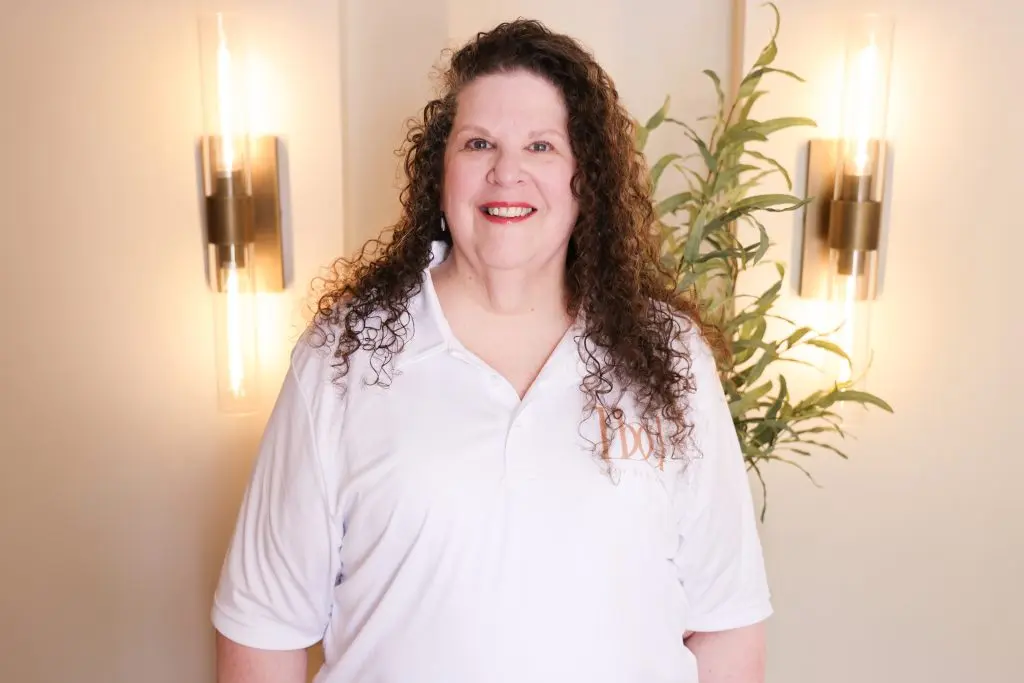 A smiling woman with curly hair, wearing a white polo shirt, stands in front of two wall sconces, with greenery visible nearby.