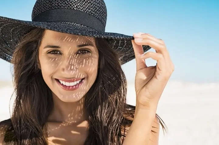 A beautiful young woman wearing summer black hat with large brim at beach. Closeup face of attractive girl with black straw hat. Happy latin woman smiling and looking at camera with sea in background.