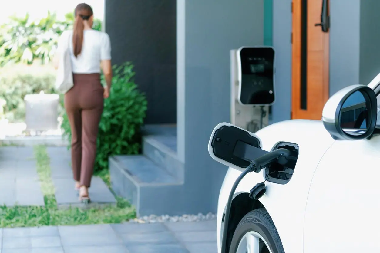 A home EV charger mounted outside a house is charging a white vehicle while a woman walks into the house