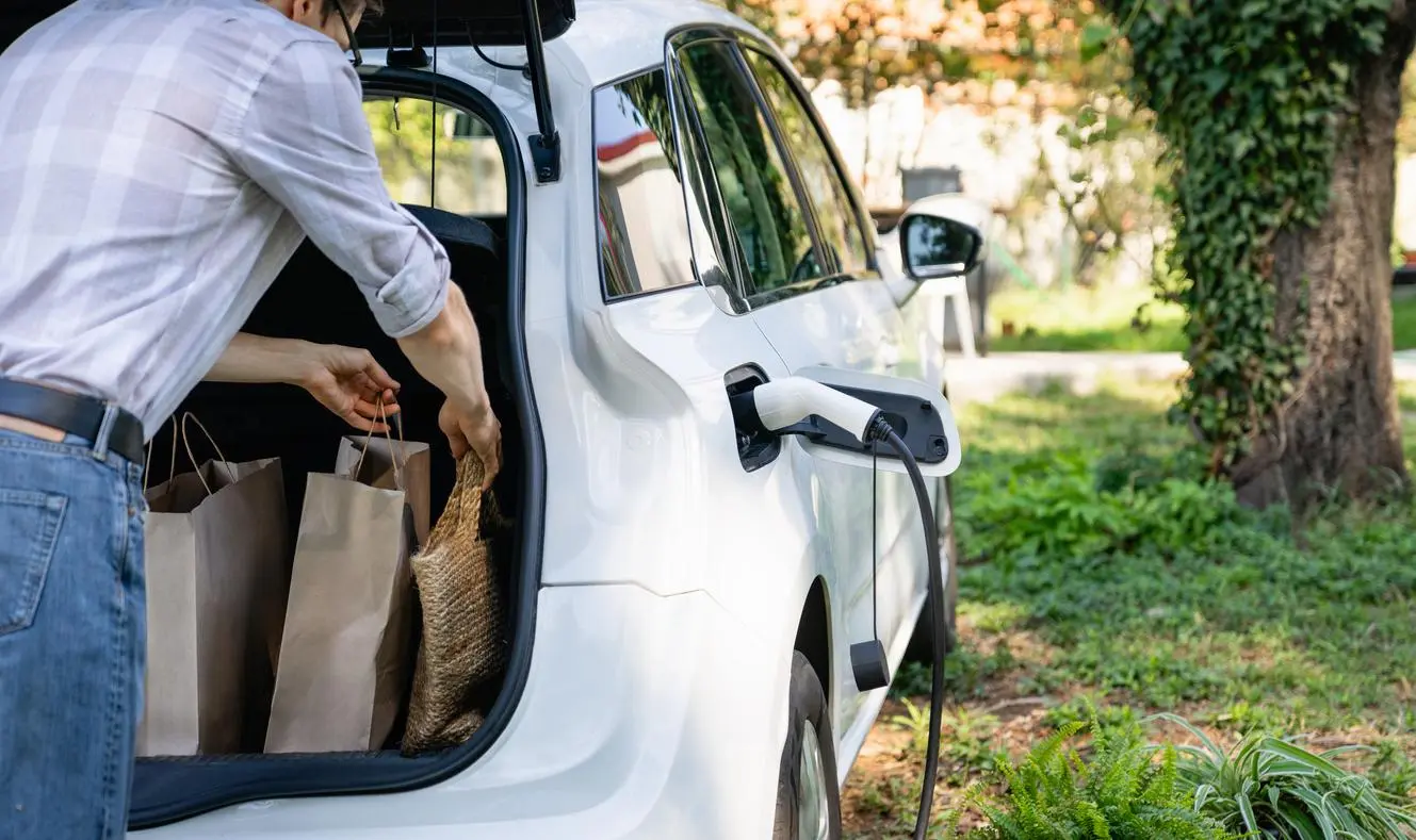 White vehicle charging with a home EV charger as a man unloads groceries from the trunk
