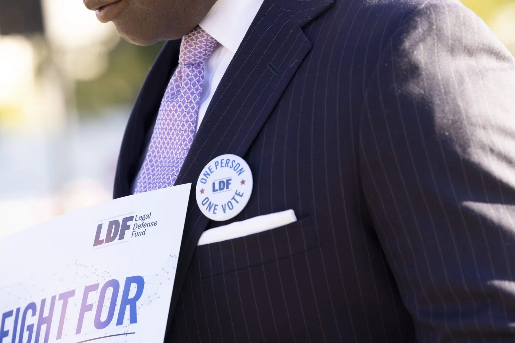 A man stands outside the Supreme Court with a sign that reads "Fight For Fair Maps" and a button that says "One Person, One Vote."