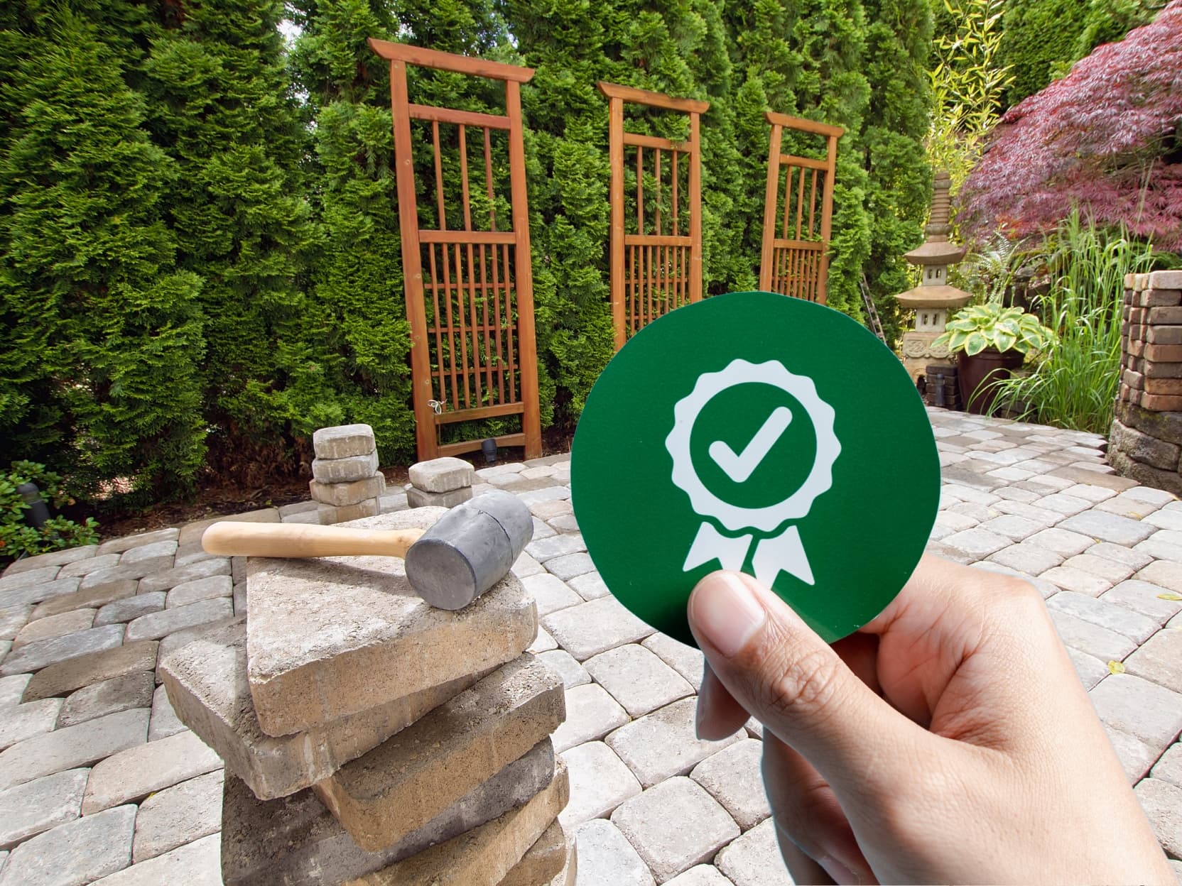 Hand holding a warranty badge symbol in front of a paver patio under construction