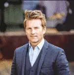 A man with light hair wearing a navy suit and vest over a light shirt is standing outdoors. The background is softly blurred, suggesting an urban or park setting.