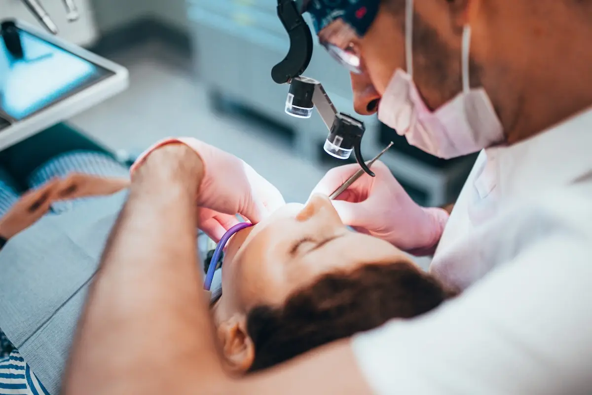 A dentist wearing a mask and magnifying glasses examines a patient's mouth, using dental instruments during a procedure. The patient is lying back in the dental chair.