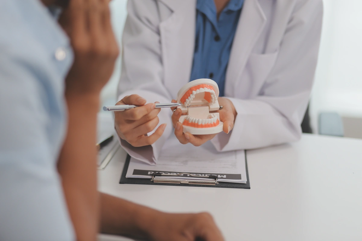 A dentist in a crisp white coat holds a dental model, using a pen to explain how long dental bonding lasts to an attentive patient. The patient listens closely, hand near their mouth, while a clipboard with important documents rests on the desk.