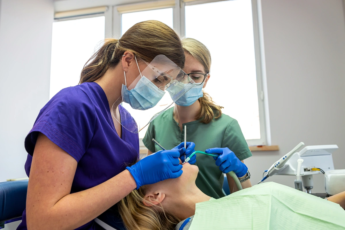 Two dentists in scrubs and face masks work on a patient reclining in the dental chair, performing a procedure like dental bonding that can last several years. One dentist holds tools, while the other assists. The bright room features a large window in the background.