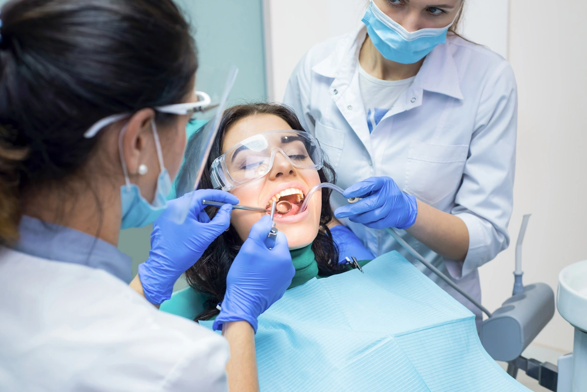 A dental procedure in progress: A patient in protective eyewear is seated in a dentist's chair, while two dental professionals, clad in blue gloves and face masks, perform an examination or treatment involving composite bonding. Dental tools and equipment are visible.